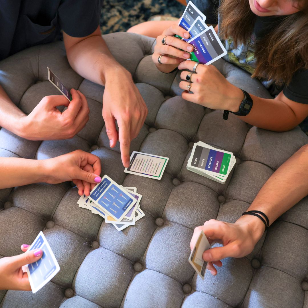 4 young adults playing Rock Paper Scissors Wizard Croc around an ottoman