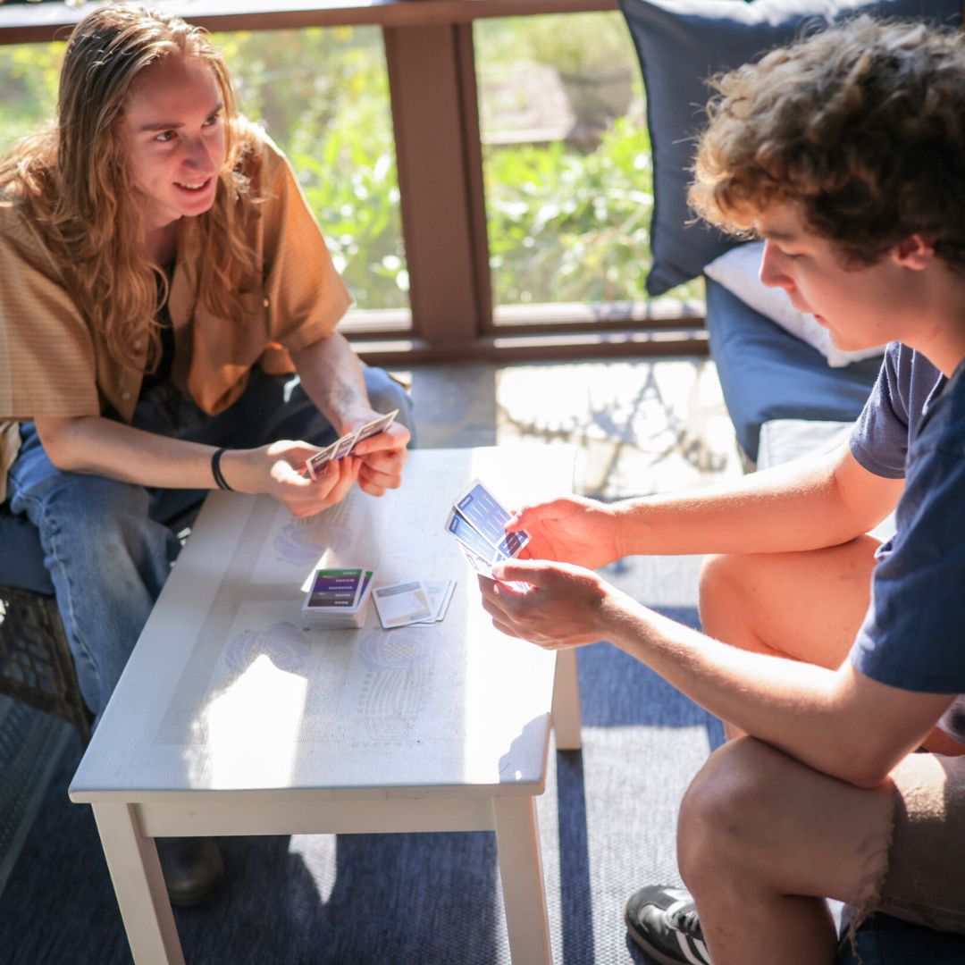 2 college guys playing Rock Paper Scissors Wizard Croc on a  porch. 1 deep in thought, the other smiling at his next move.