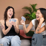2 women laughing out loud while playing Stack the Deck on a couch in front of a large, green plant