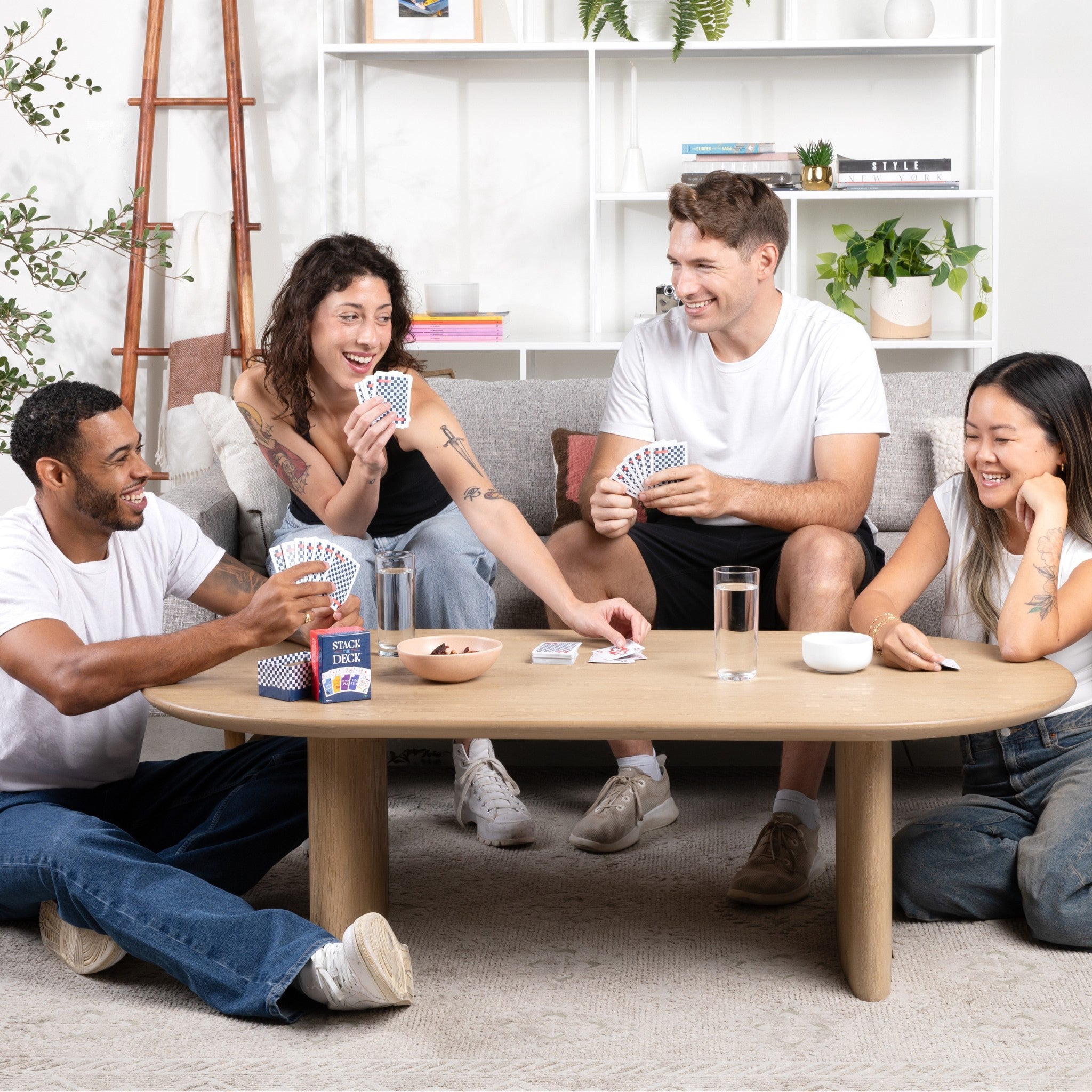 4 young adults laughing and playing Stack the Deck in a living room setting with cards and snacks