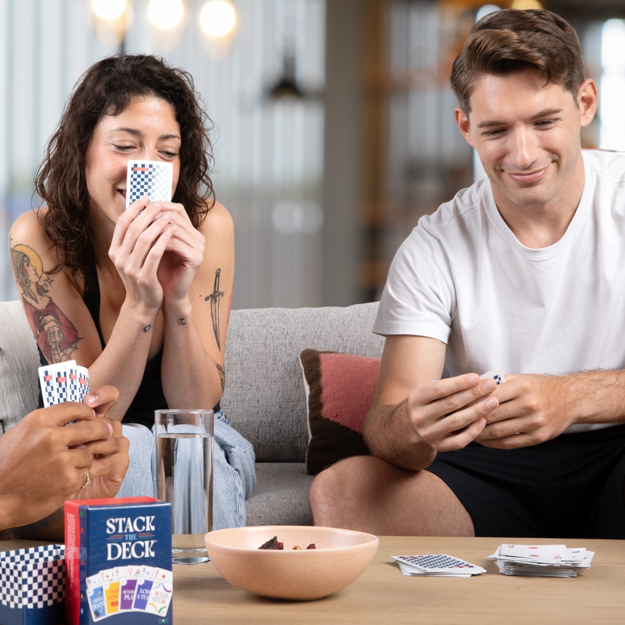 People gathered on a couch play Stack the Deck, smiling as cards, snacks, and a drink sit on the coffee table.