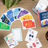 Playing cards with colorful text and graphics on a wooden table with a 'Stack the Deck' box and plant.