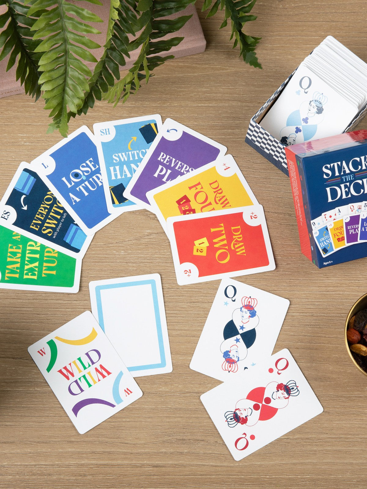 Playing cards with colorful text and graphics on a wooden table with a 'Stack the Deck' box and plant.