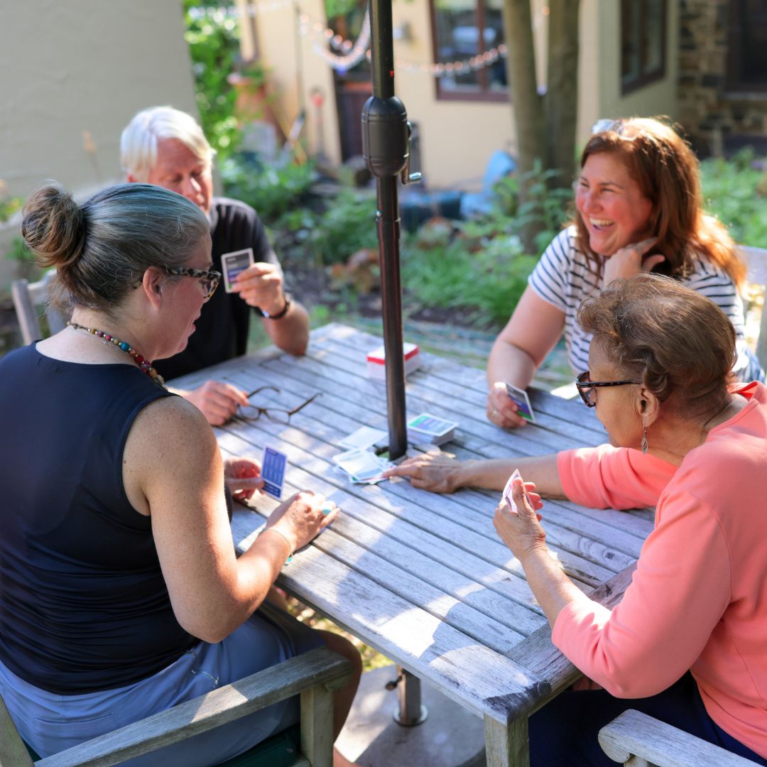 Group of 4 older adult friends laughing while playing cards outdoors at a wooden table with umbrella.