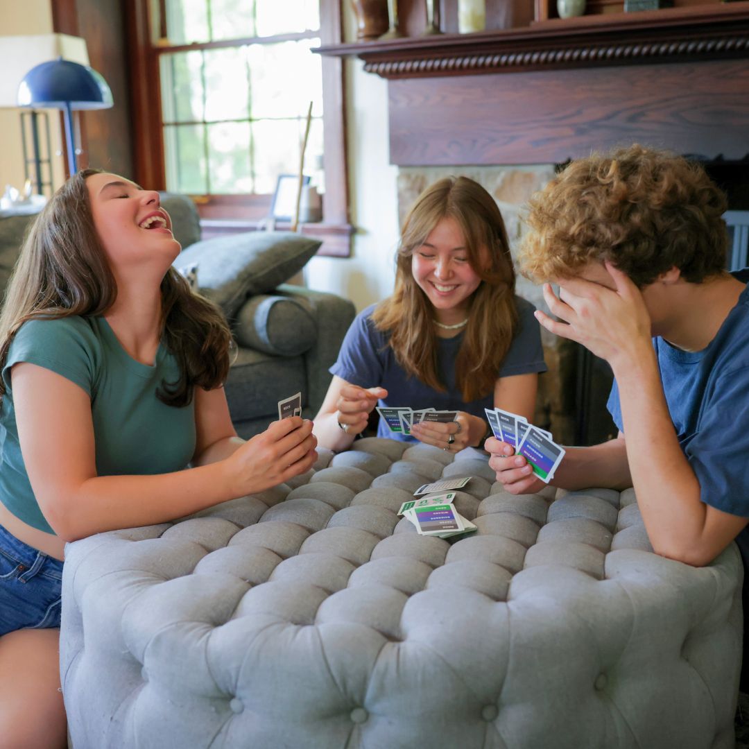 3 high-school and college-aged students laughing wildly while playing Rock Paper Scissors Wizard Croc in a family room..
